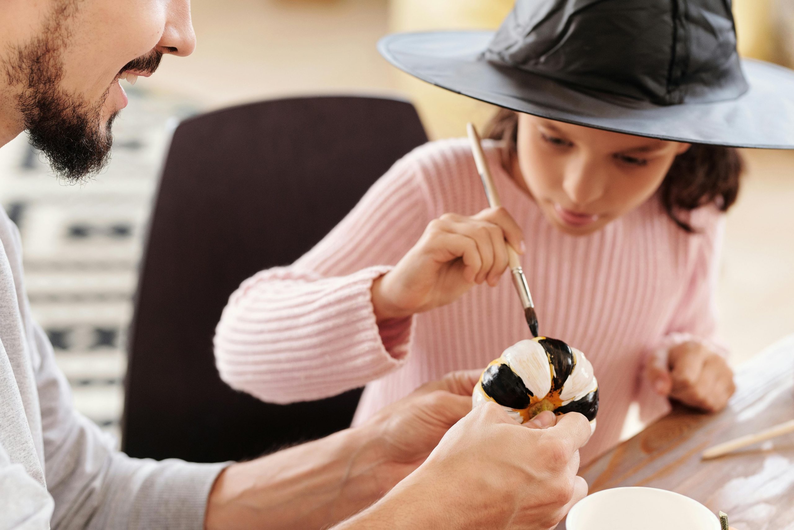 A child wearing a witch's hat painting a pumpkin. The pumpkin is being steadied by her father.