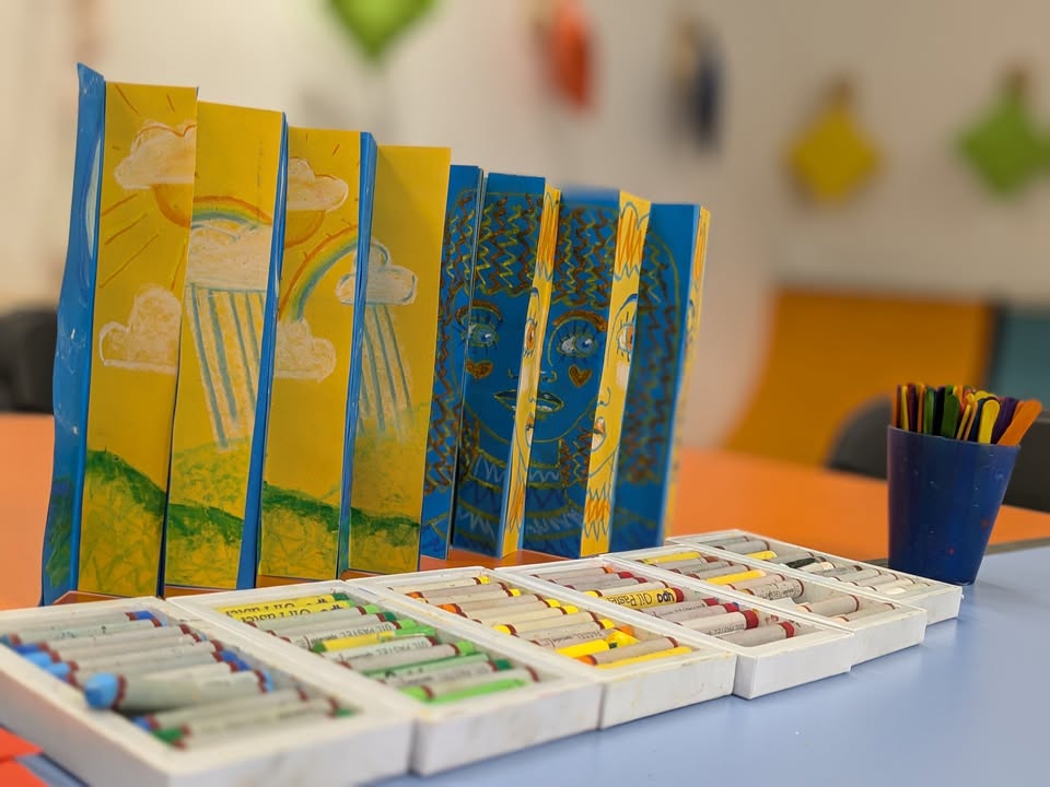 A blue and yellow artwork stands on a blue table, in front of it are boxes of oil pastels. In the background there is a dark blue cup of multicoloured