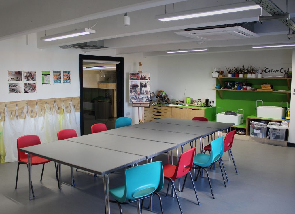 Wide shot showing the Learning Studio. There is a table in the middle of the room surrounded by 8 chairs and a lrage sink on the back wall with arts and crafts equipment around it.