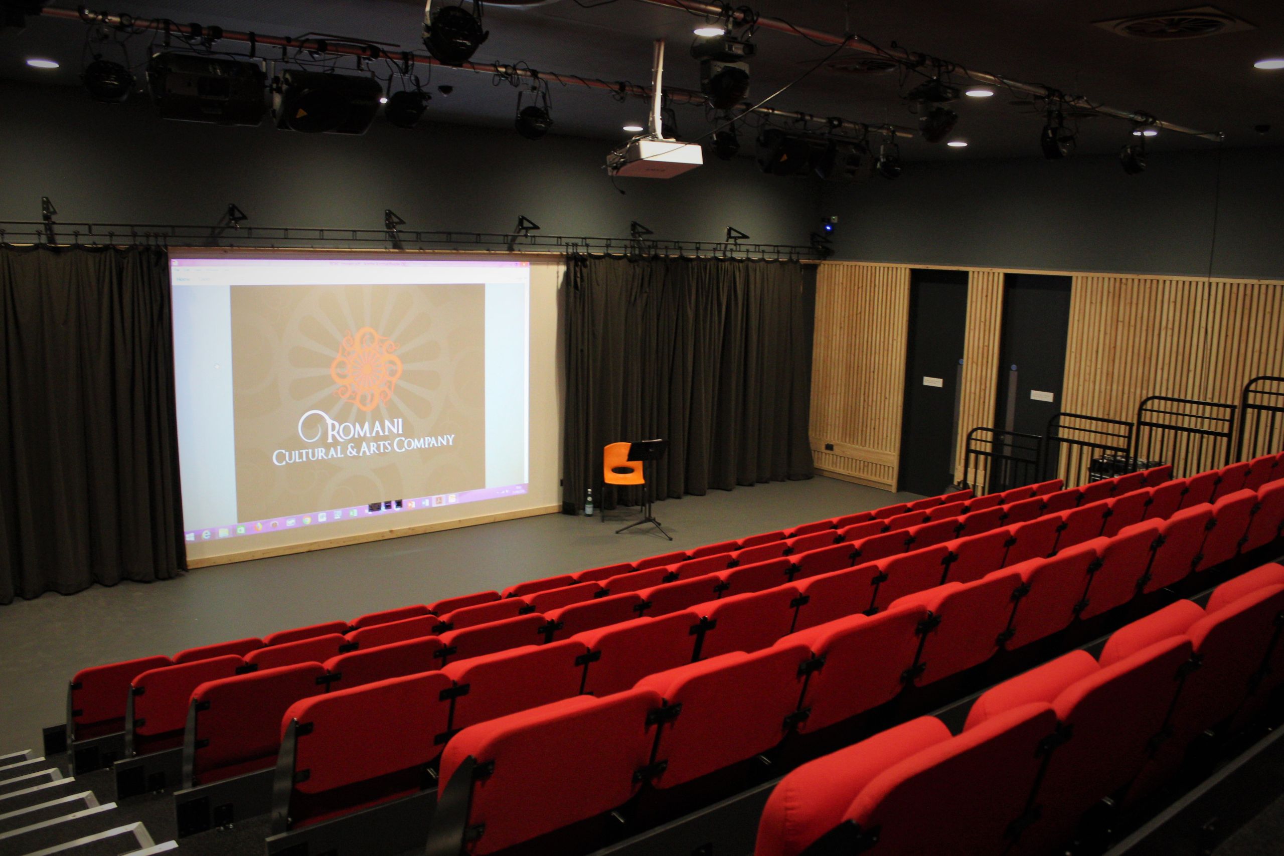 Wide shot of the performance space, taken from the back of the room, showing the red seats and the projector screen at the front.